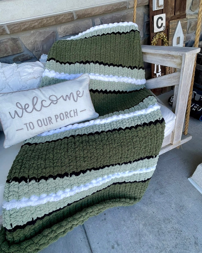 Green striped blanket draped over a chair with a 'Welcome to our porch' pillow on a stone porch.